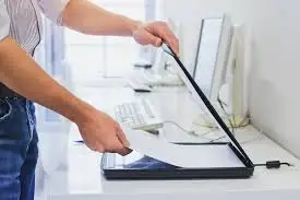 Persona colocando una hoja de papel en un escáner de superficie plana en una oficina con computadoras en el fondo.